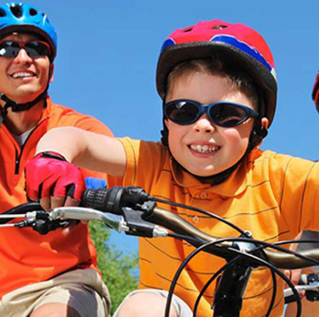 A smiling child and adult riding mountain bikes, wearing helmets and sunglasses on a sunny day